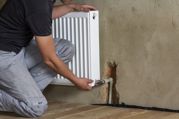 Professional plumber worker installing heating radiator on brick wall in an empty room of a newly built apartment or house.